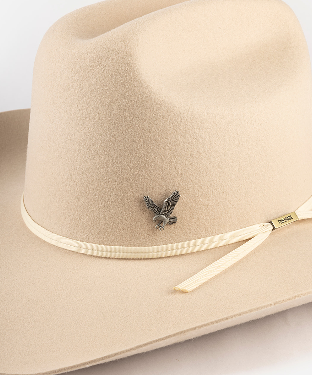 A close-up of a beige felt cowboy hat featuring a small silver eagle pin on the side and a matching beige ribbon with a gold-toned tip #color_anti silver
