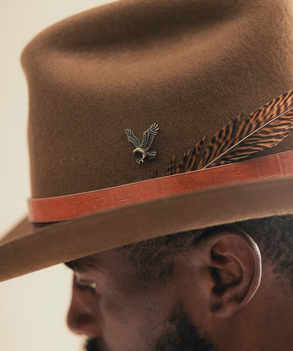 Close-up of a brown felt cowboy hat worn by a man, featuring a reddish-brown band, a feather accent, and a small eagle pin #color_anti silver