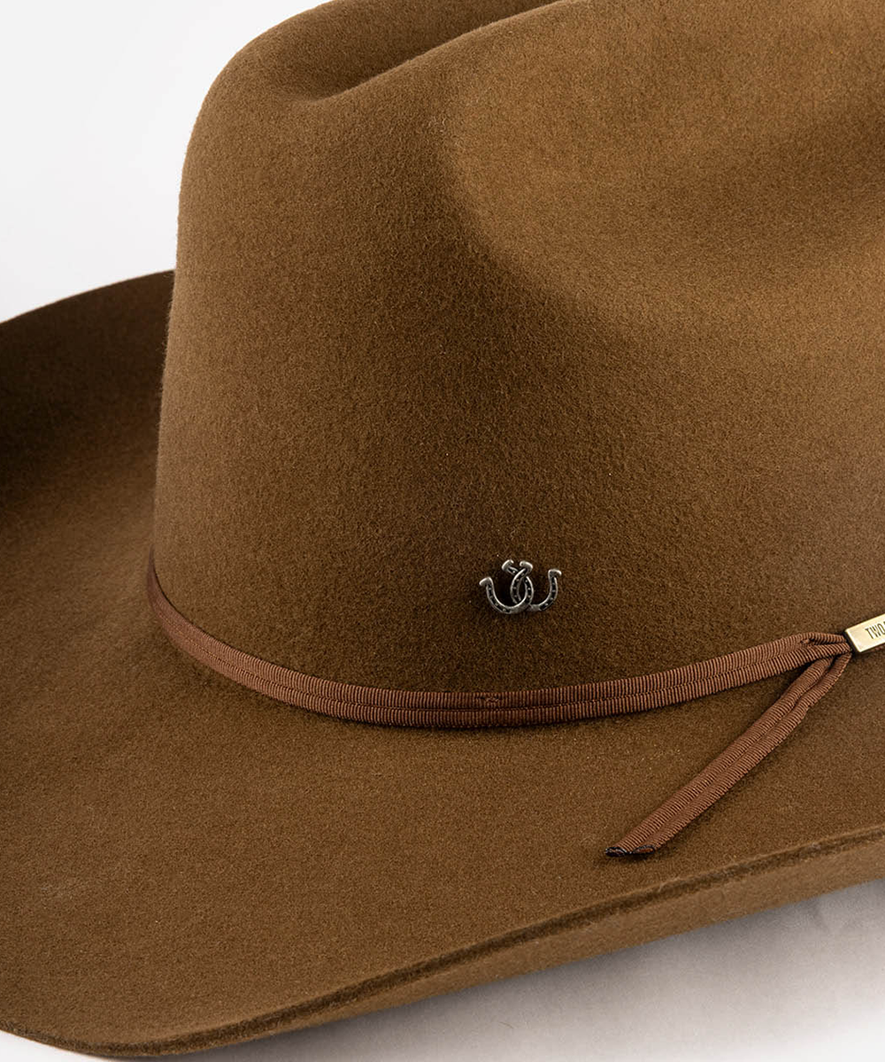 A close-up of a brown felt cowboy hat featuring a matching brown ribbon band with a small gold-toned decorative emblem and a small white brand tag #color_anti silver
