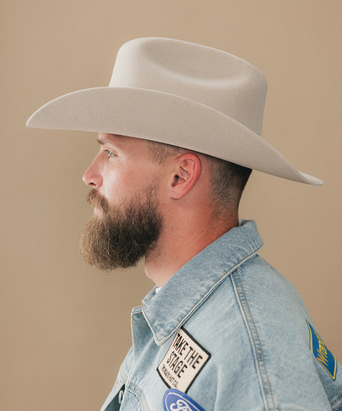 A man with a full beard wearing a cream felt cowboy hat and a denim jacket, shown in a side profile against a solid beige background #color_silverbelly