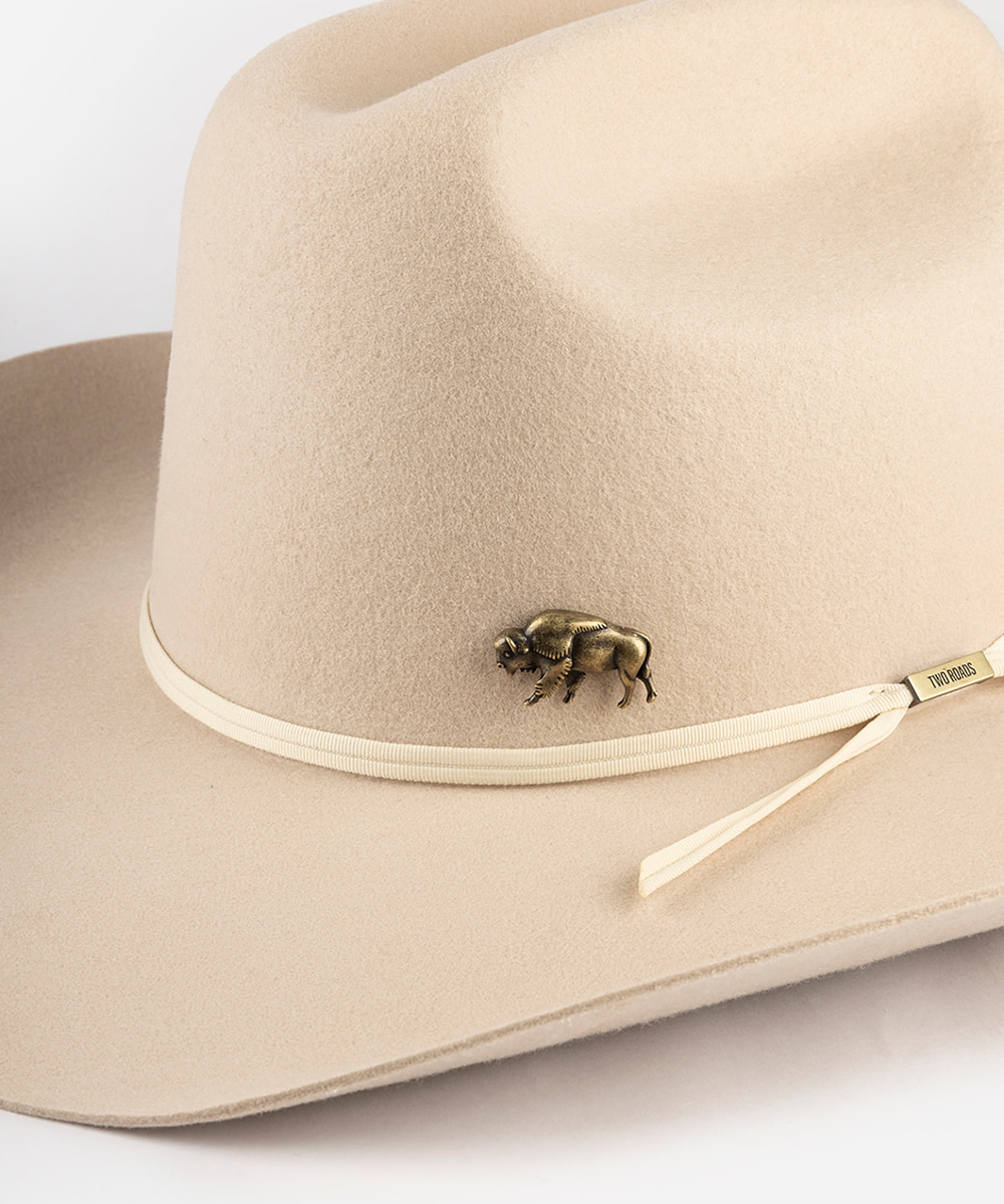 A close-up of a beige felt cowboy hat featuring a small bronze bison pin on the side of the crown and a matching beige ribbon with a small gold-toned tag #color_anti gold