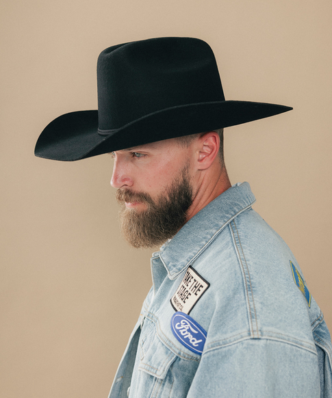 A man with a full beard wearing a black felt cowboy hat and a denim jacket, shown in a side profile against a solid beige background #color_black