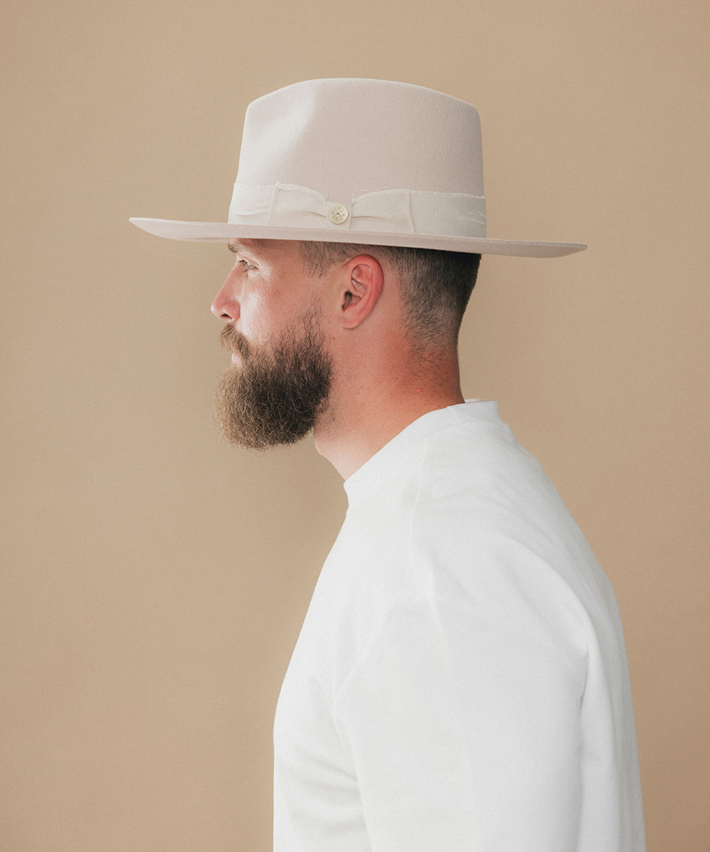 A man with a full beard wearing a cream felt fedora hat and a white t-shirt, shown in a side profile against a solid beige background #color_ivory