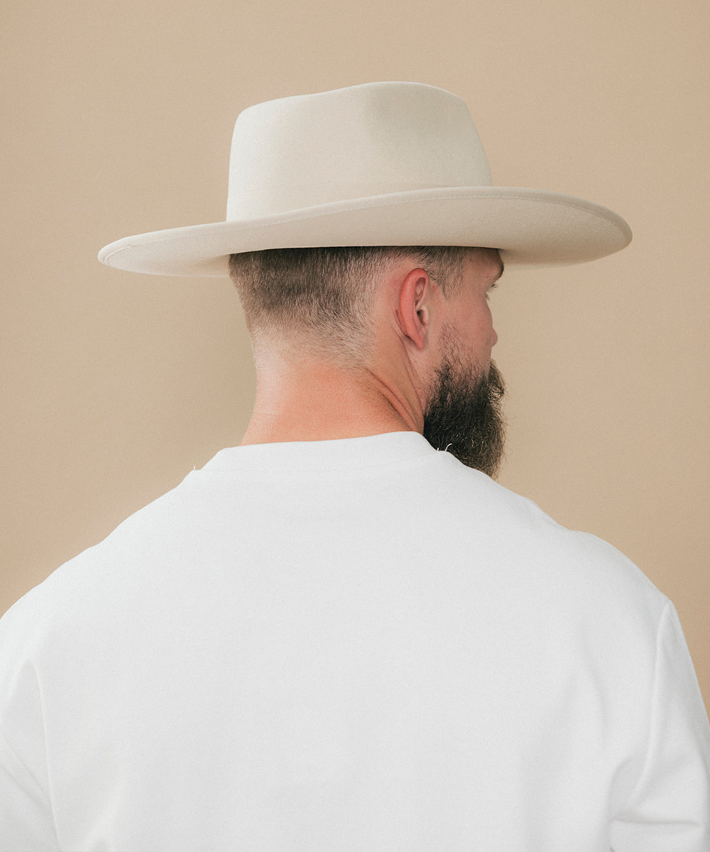 A man shown from the back wearing a light beige wide-brim fedora hat and a white t-shirt, set against a solid beige background #color_buckskin