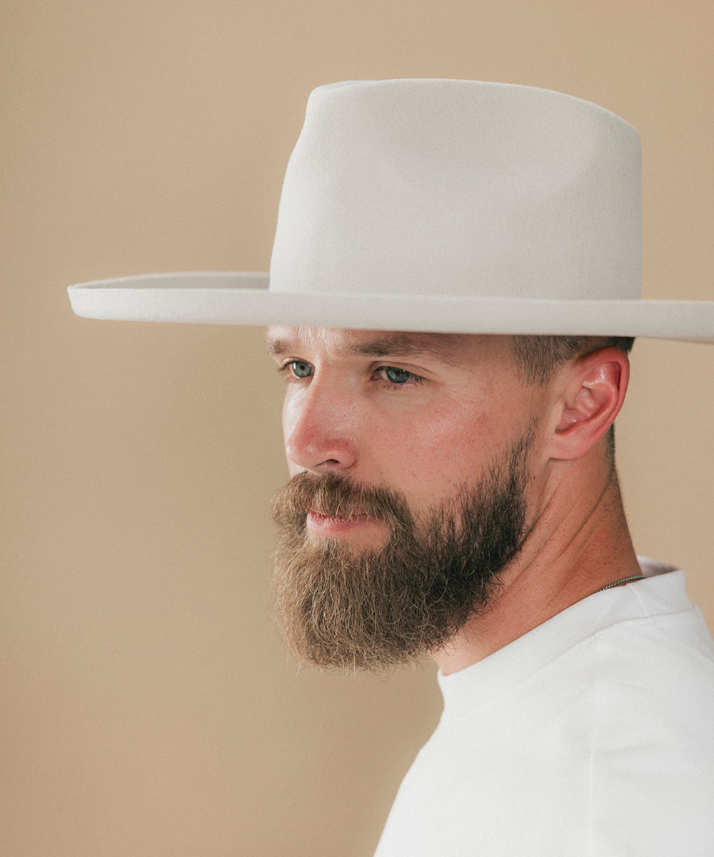 A man with a full beard wearing a white felt wide-brim fedora hat and a white t-shirt, shown in a side profile against a solid beige background #color_silver grey
