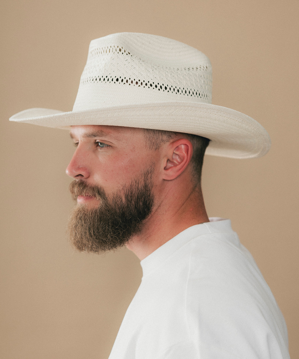 A man in profile wearing a white woven cowboy hat and a white shirt, photographed against a plain warm beige studio background #color_ivory
