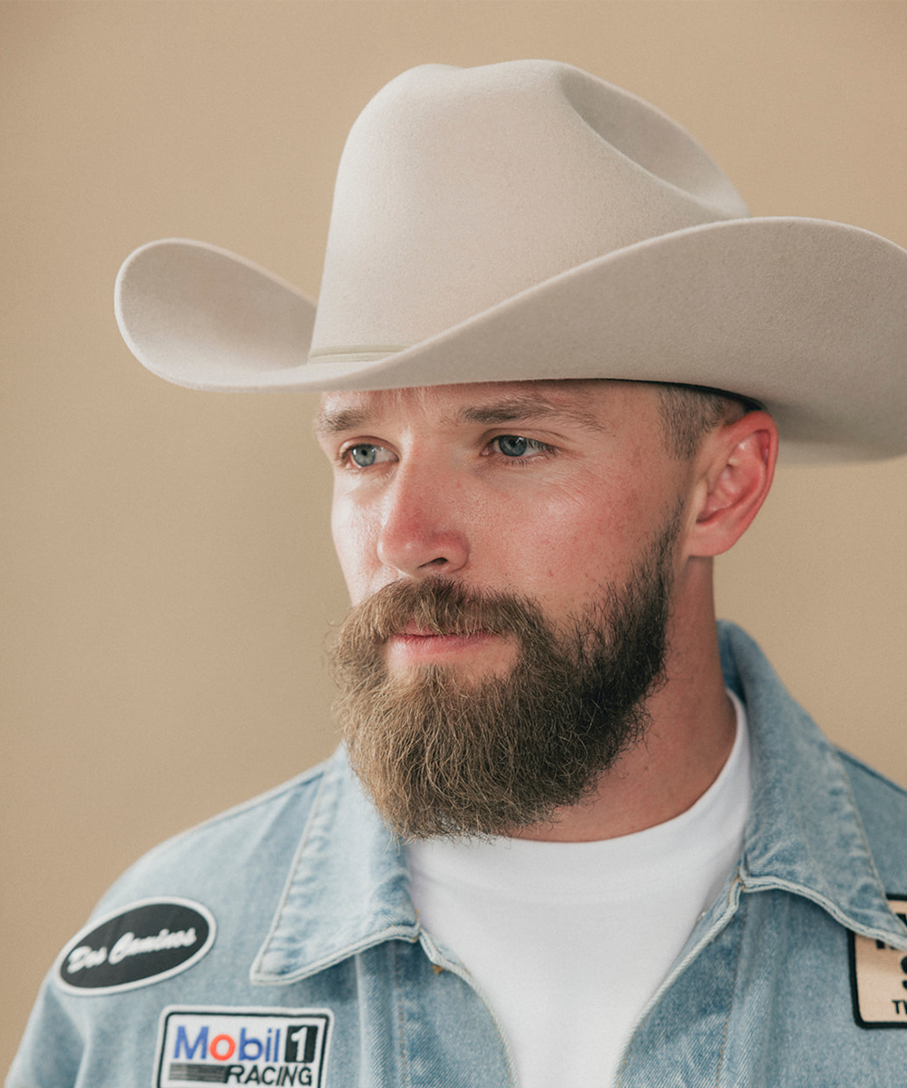 A close-up portrait of a man wearing a light beige cowboy hat and a light blue denim racing shirt with Mobil 1 Racing patches, set against a solid beige background #color_silverbelly