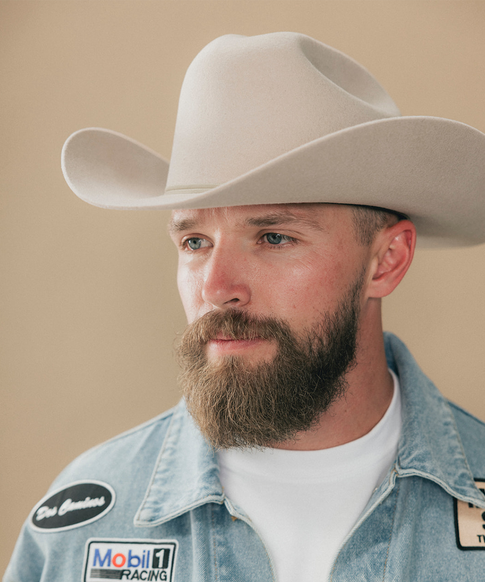 A close-up portrait of a man wearing a light beige cowboy hat and a light blue denim racing shirt with Mobil 1 Racing patches, set against a solid beige background #color_silverbelly