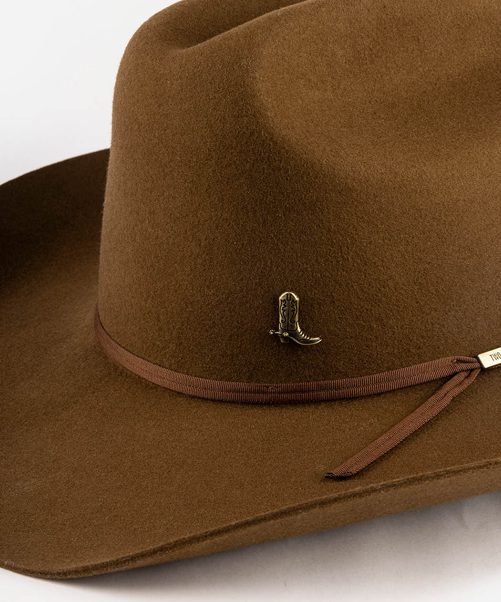 A close-up of a brown felt cowboy hat featuring a matching brown ribbon band with a small gold-toned boot-shaped pin and a white brand tag #color_anti gold