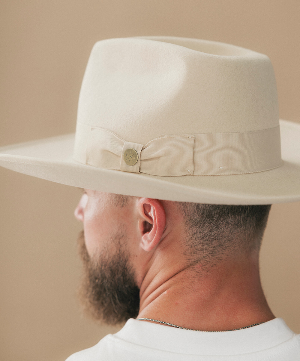 A man shown from the side and back wearing a cream-colored fedora hat with a matching band and bow detail, set against a solid beige background #color_buckskin
