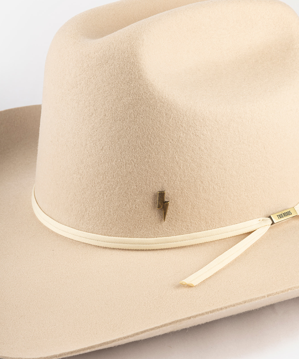 A close-up of a beige felt cowboy hat with a cream-colored band, a small gold lightning bolt pin, and a gold brand tag #color_anti gold