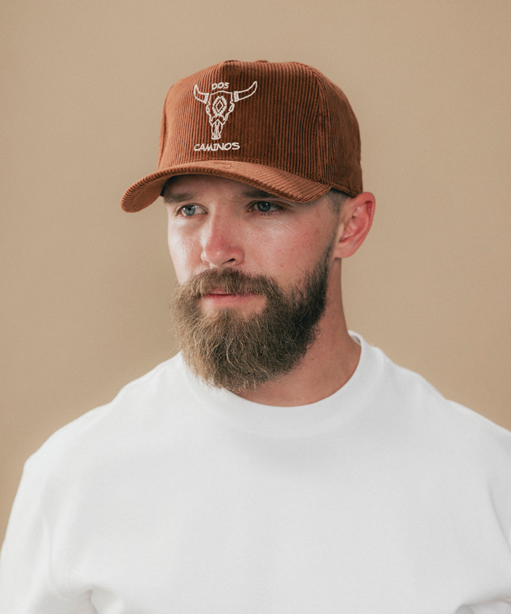 A man with a full beard wearing a brown corduroy baseball cap with a white embroidered bull skull logo and the brand name 'DOS CAMINOS' on the front, paired with a white crewneck t-shirt against a solid beige background #color_sienna