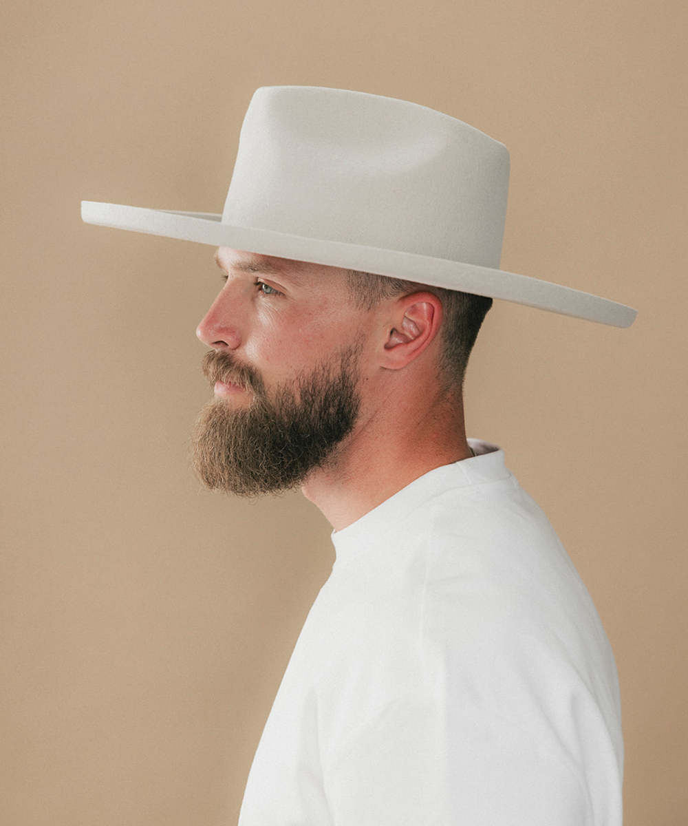 A man with a full beard wearing a white felt wide-brim fedora hat and a white t-shirt, shown in a side profile against a solid beige background #color_silver grey