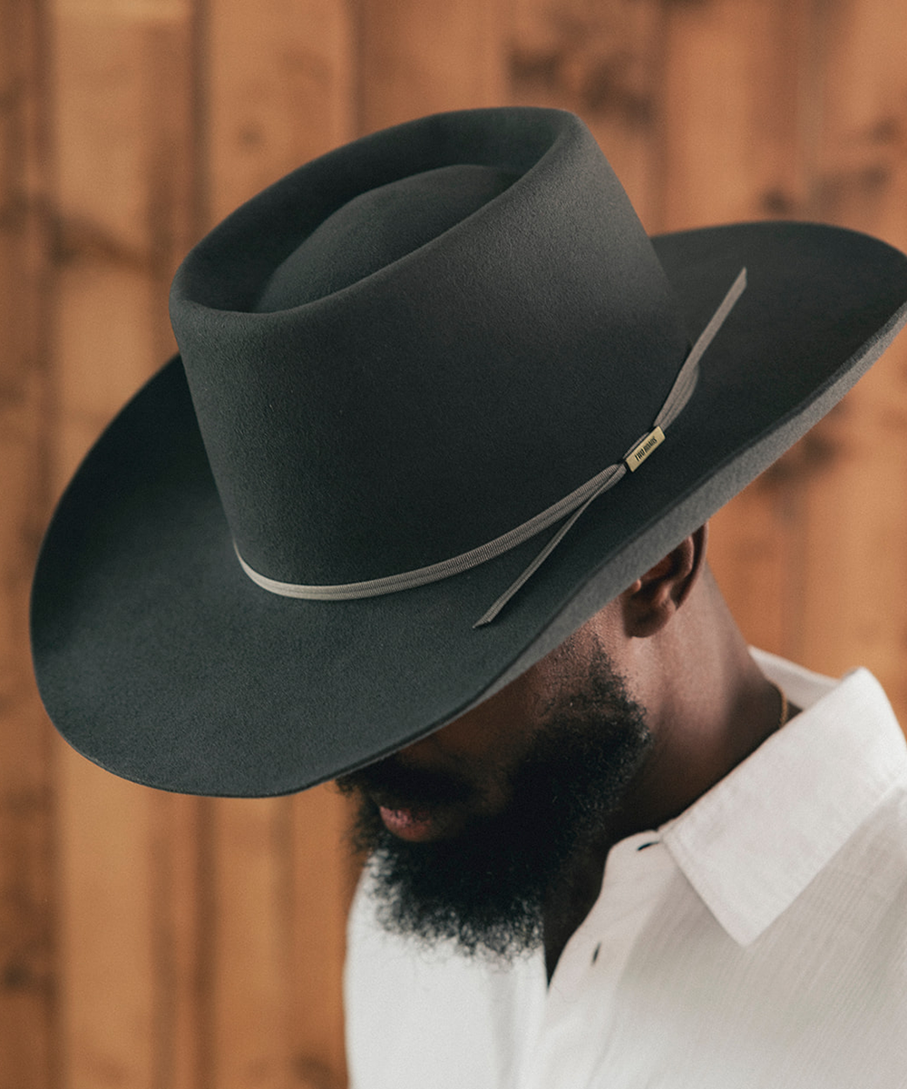 A man wearing a wide-brimmed black cowboy hat with a thin gray band and a small gold accent, photographed against a warm wood plank background #color_grey