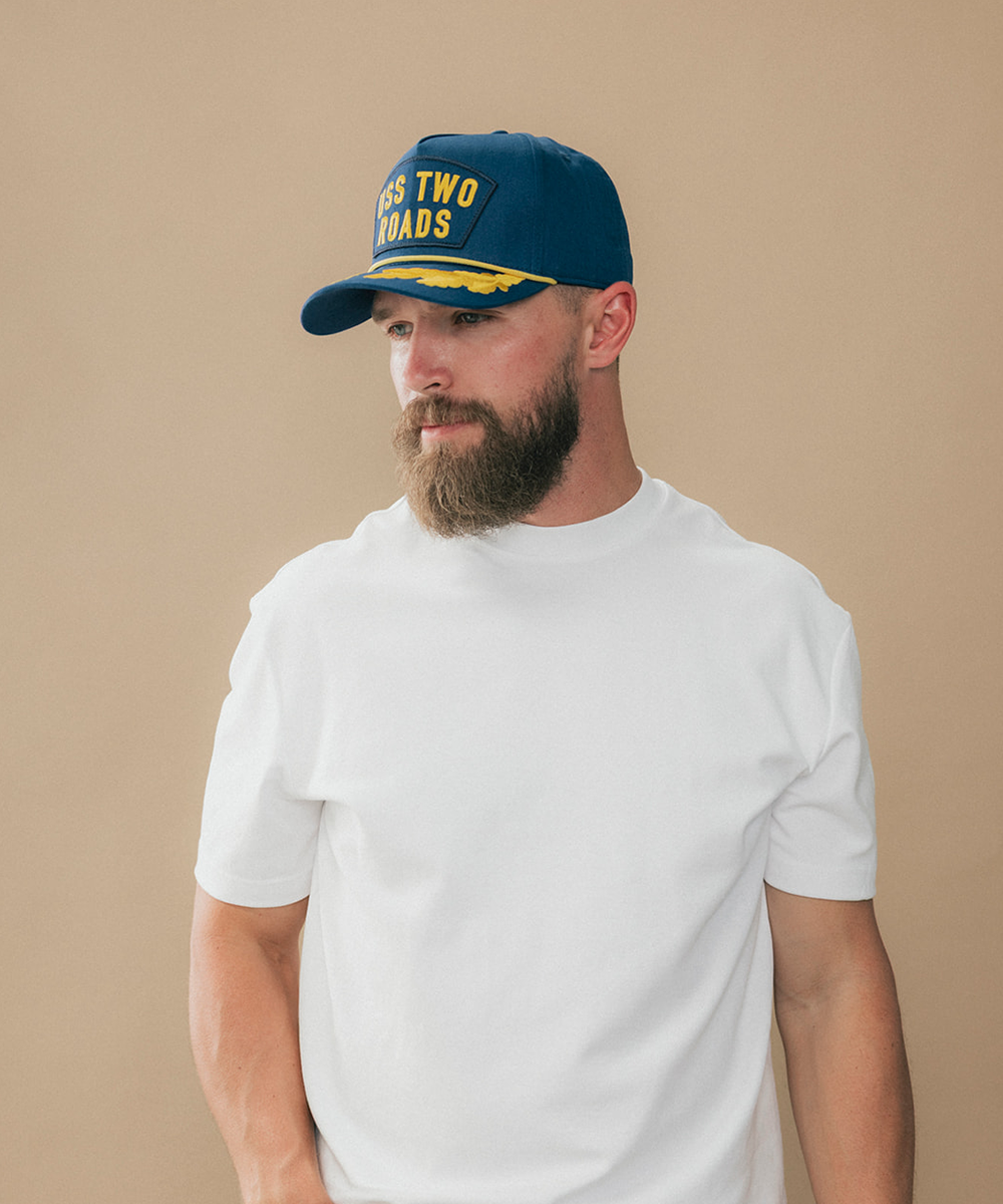 A man wearing a navy blue baseball cap with yellow embroidered text reading 'TWO ROADS' and a yellow stripe detail, paired with a plain white t-shirt, set against a solid beige background #color_blue
