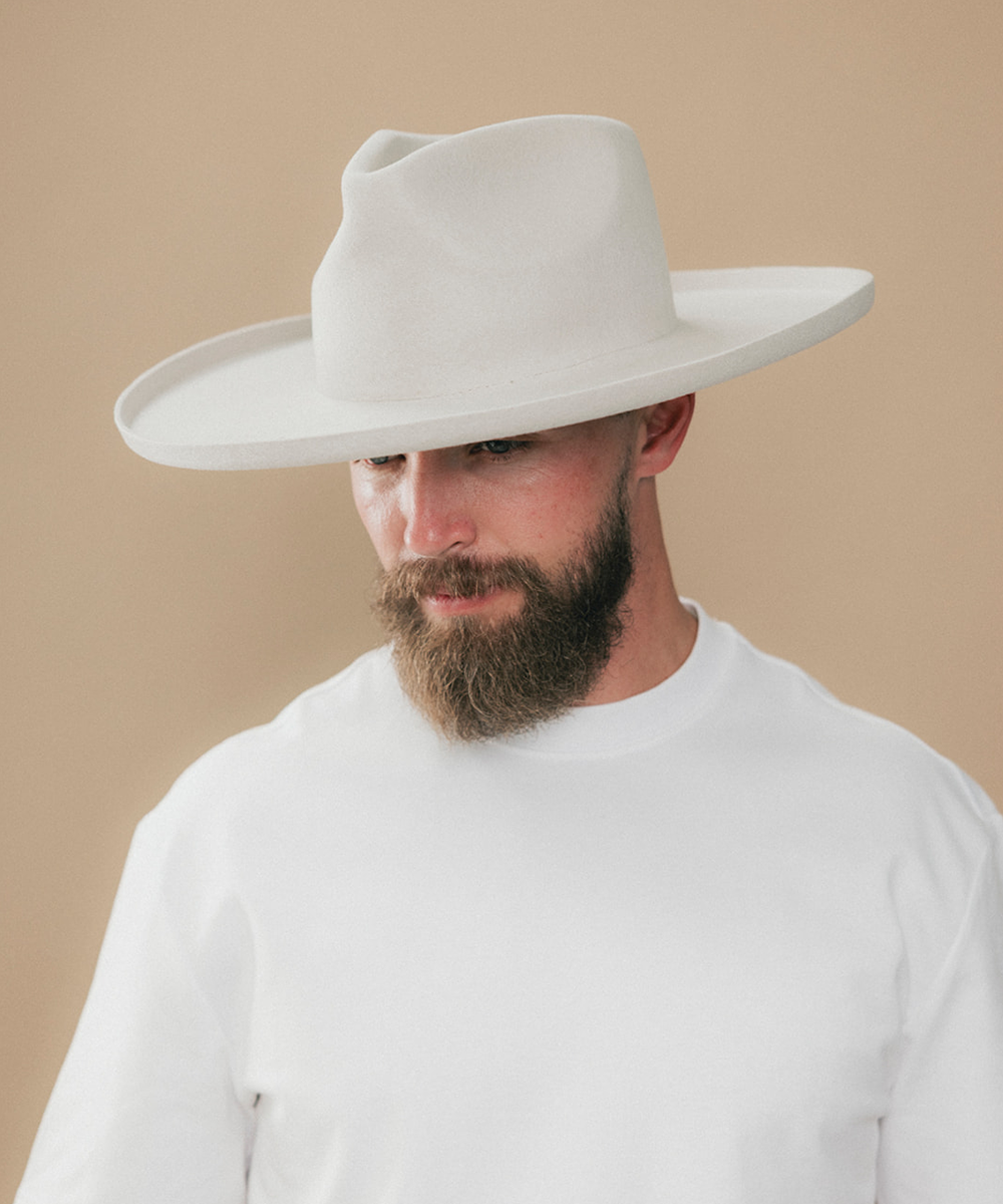 A man with a full beard wearing a white felt wide-brim fedora hat and a white t-shirt, shown in a side profile against a solid beige background #color_silver grey