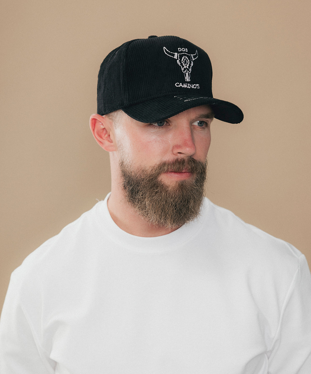 A man with a beard wearing a black baseball cap with a white bull skull logo and the text 'DOS CAMINOS' on the front, paired with a white crewneck t-shirt against a solid beige background #color_obsidian