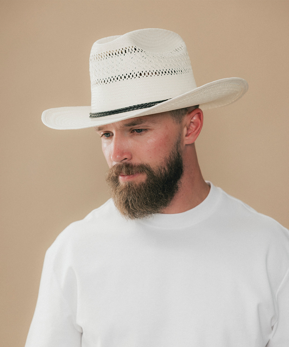 A man with a full beard wearing a white woven cowboy hat with a black band and a white t-shirt, photographed against a solid beige background #color_ivory