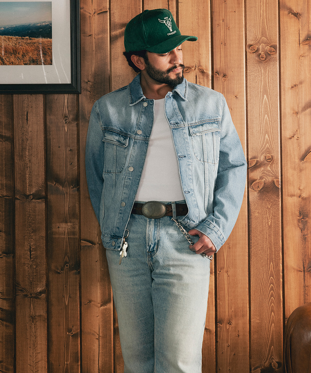 Man wearing a denim jacket and baseball hat standing against a wooden wall #color_saguaro