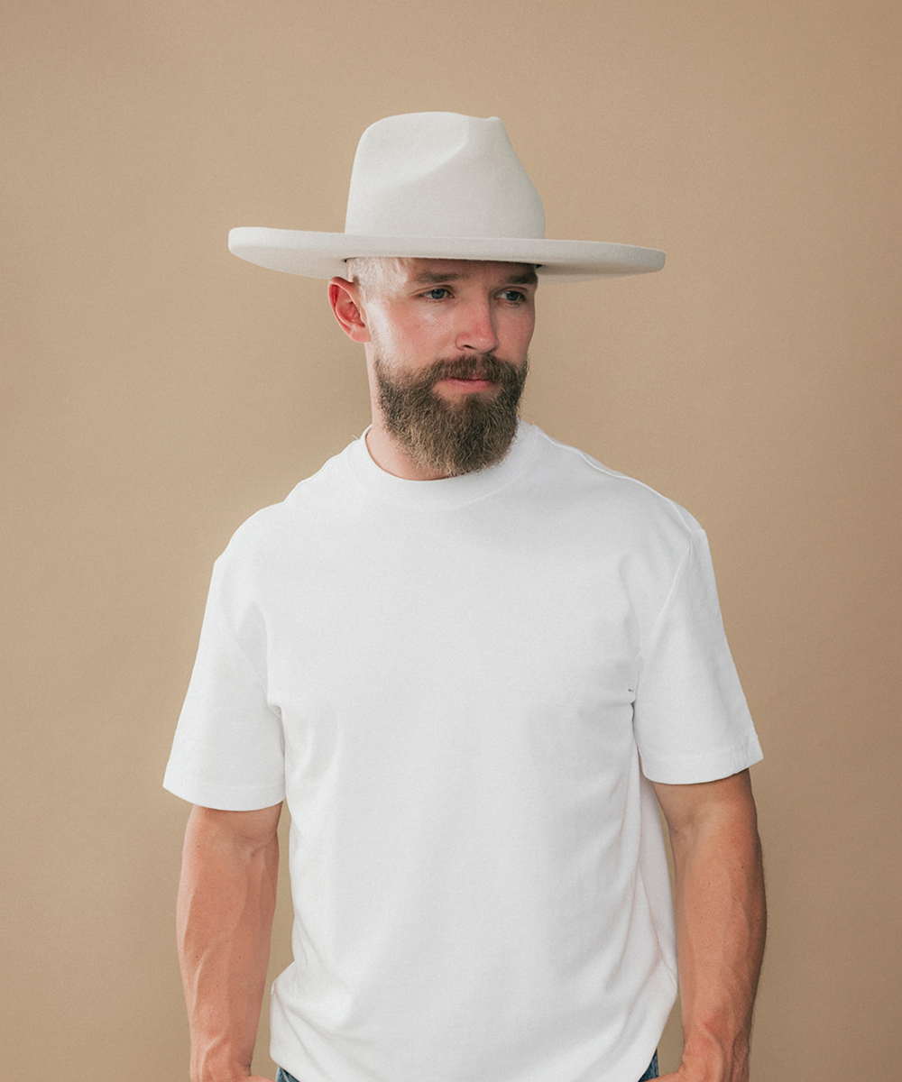 A man with a full beard wearing a white felt wide-brim fedora hat and a white t-shirt, shown in a side profile against a solid beige background #color_silver grey