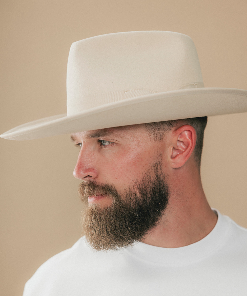 A man in profile wearing a light cream-colored cowboy hat and a white t-shirt, set against a solid beige background #color_buckskin