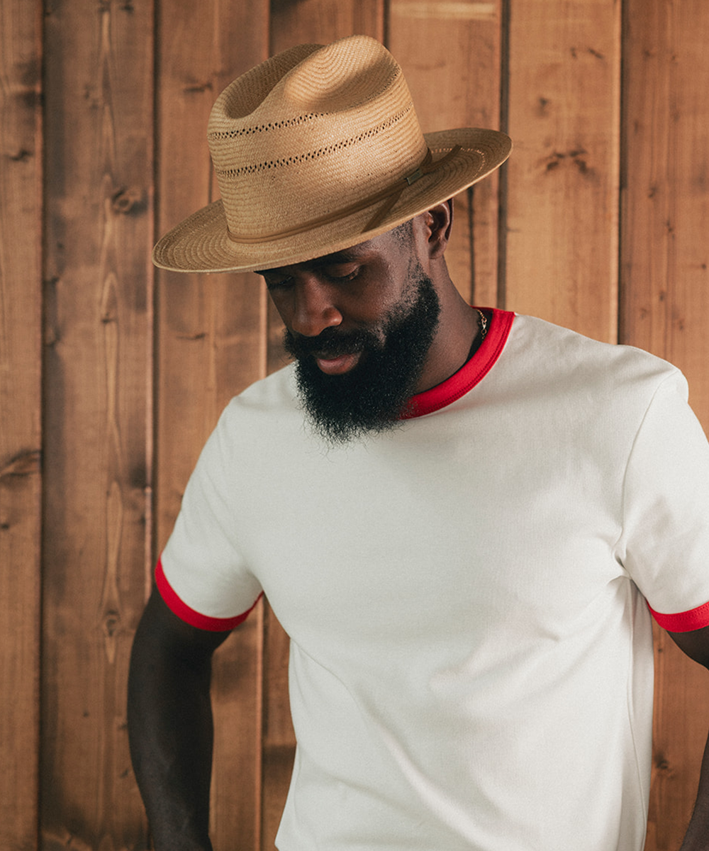 Man sitting on a stool in a room with wooden paneling, wearing a white shirt with red trim and blue jeans #color_natural