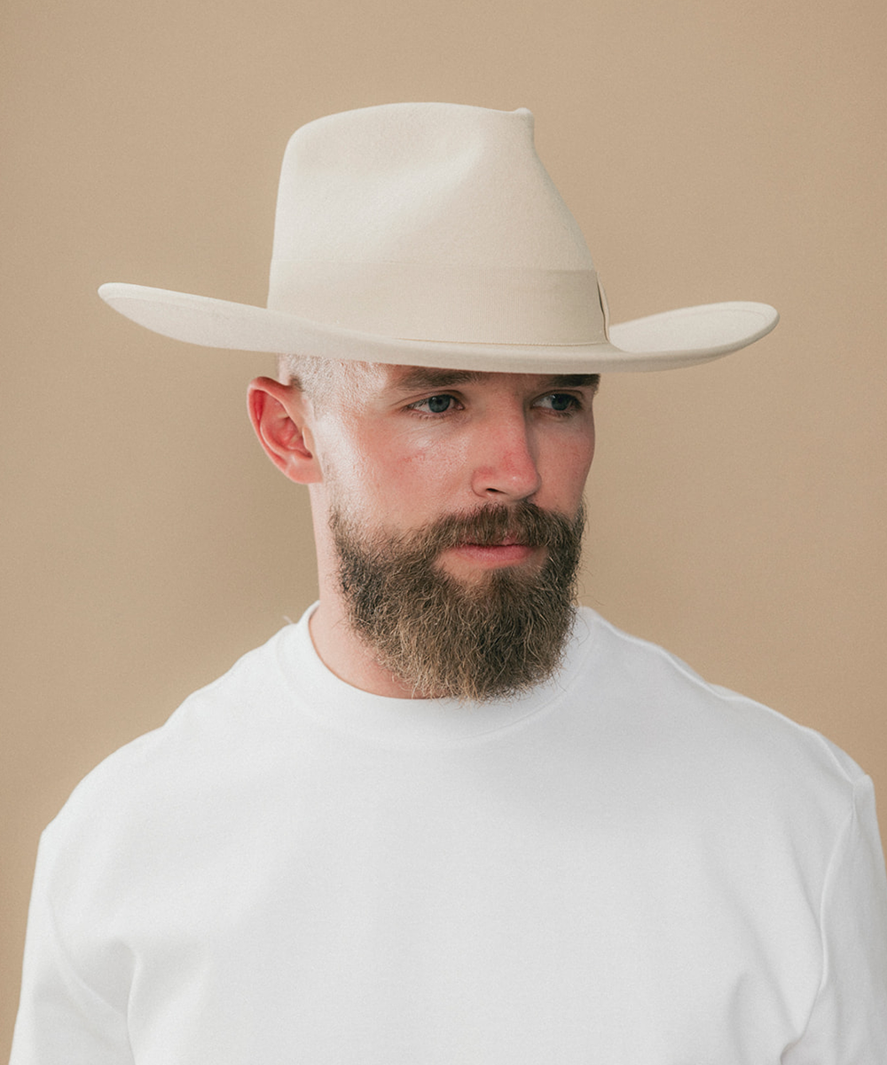 A man with a full beard wearing a light beige cowboy hat and a white t-shirt, photographed against a solid beige background #color_buckskin