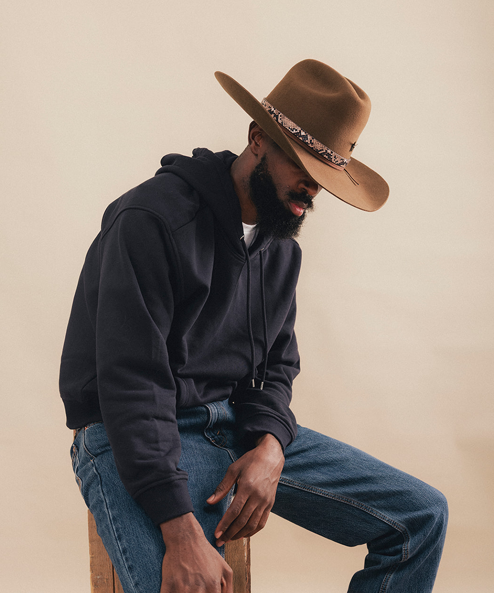 A man wearing a dark navy hoodie, blue jeans, and a brown cowboy hat with a patterned band, sitting on a wooden stool against a plain beige background #color_tobacco