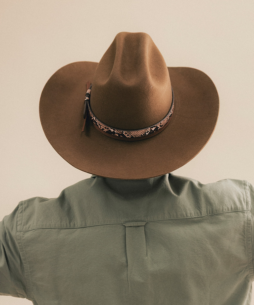 A person shown from behind wearing a brown wide-brim cowboy hat with a patterned band and an olive green shirt against a plain beige background #color_tobacco